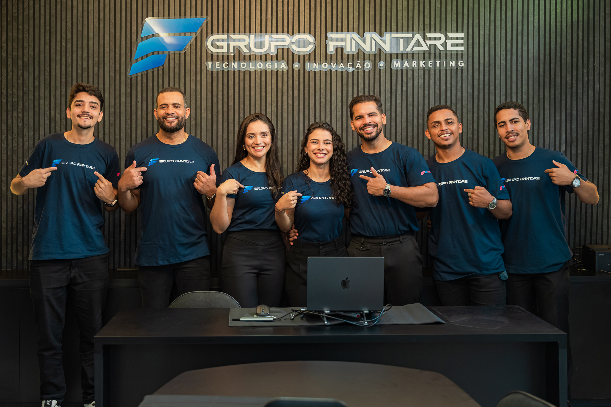 Parte do time do Grupo Finntare reunida no escritório, com camisetas da marca, diante do logo Tecnologia · Inovação · Marketing.
